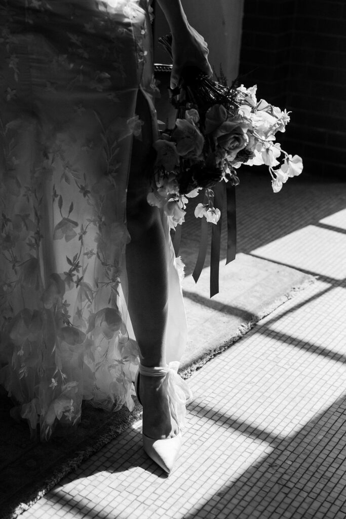 Bride stands in the natural daylight of window while holding her bridal bouquet