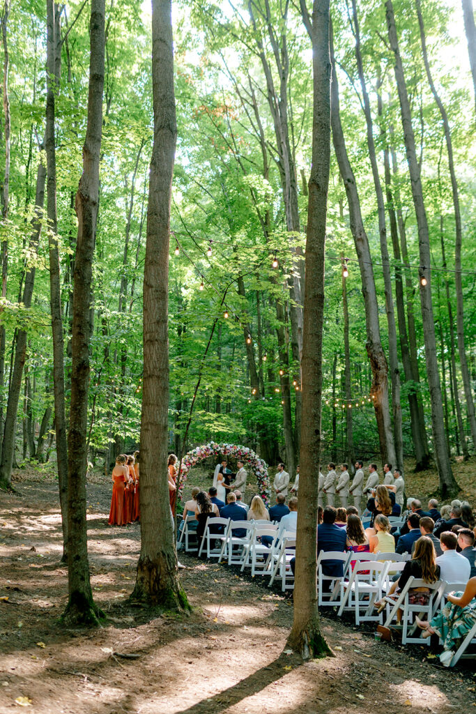 Wedding guests look on as bride and groom exchange vows under filtered light through the trees