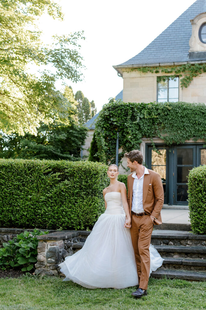 Bride and groom walk down stone steps with ivy-draped manor in the background