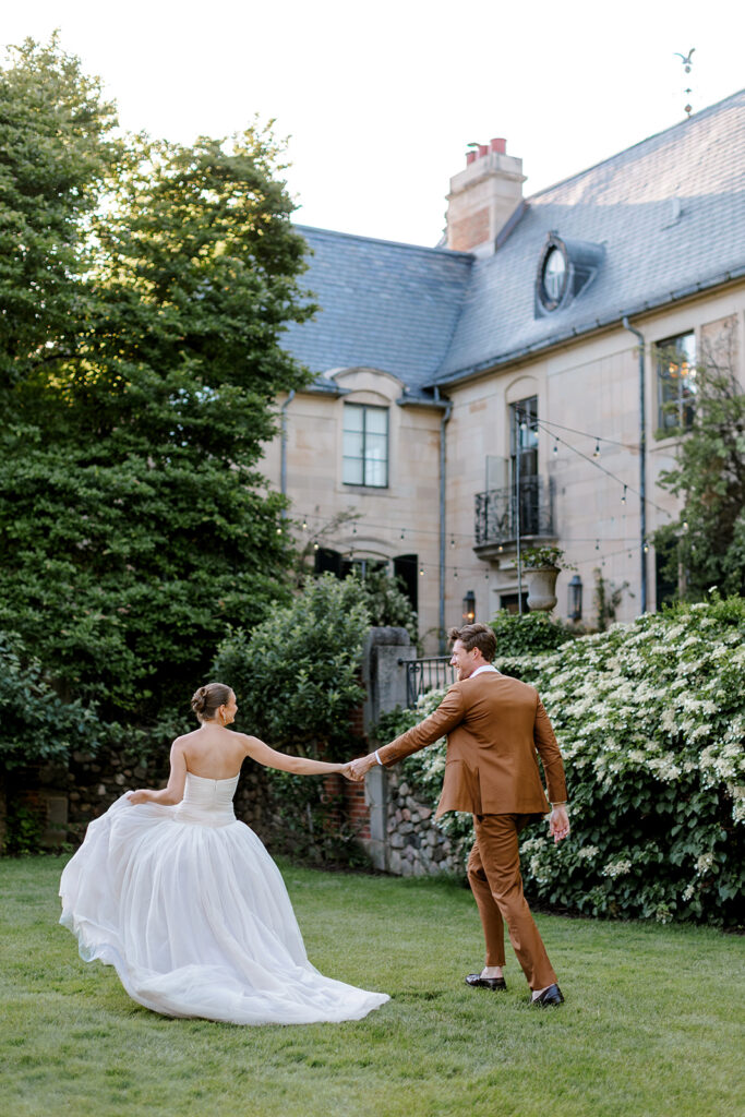 Bride and groom traipse hand in hand across lawn of European style wedding venue