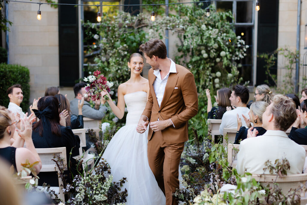 Bride smiles at groom as they walk hand in hand down the aisle to the applause of wedding guests
