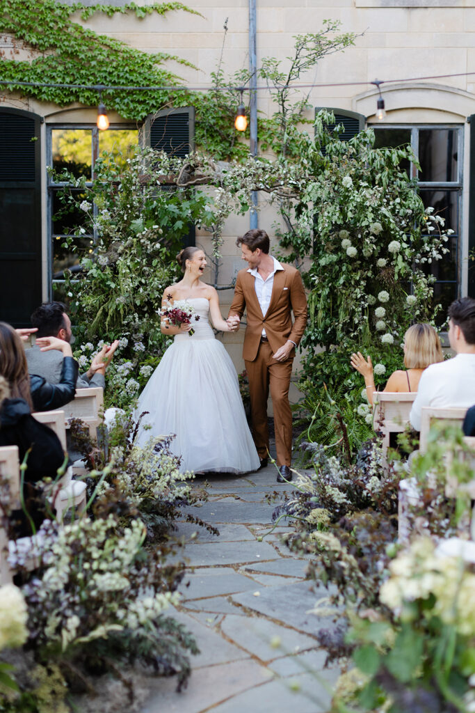 Bride and groom under ceremony arch blooming with roses and wild greenery at European style wedding venue

