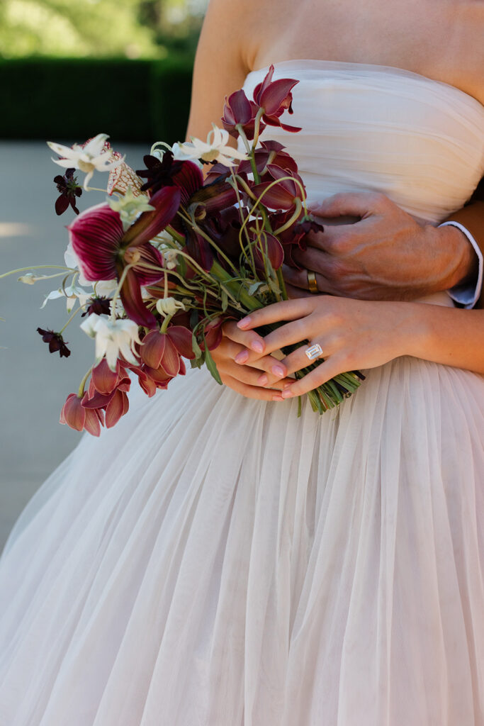 Bride holds her richly-colored bouquet while groom wraps his arms around her waist at European style wedding venue