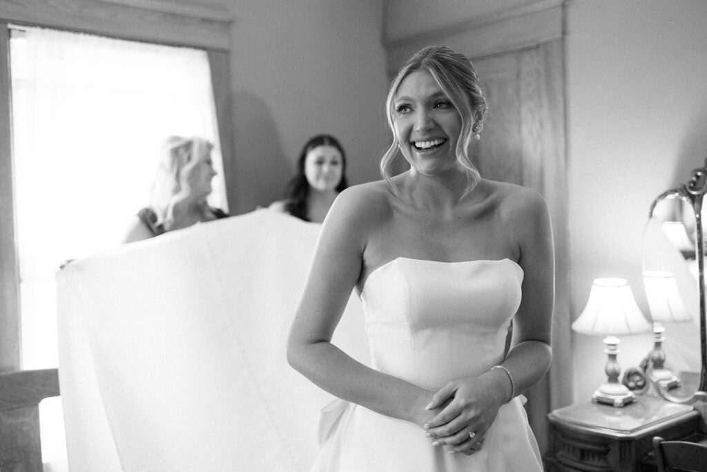 Bride smiling as bridesmaids assist with her dress, a joyful behind-the-scenes moment for your wedding shot list.