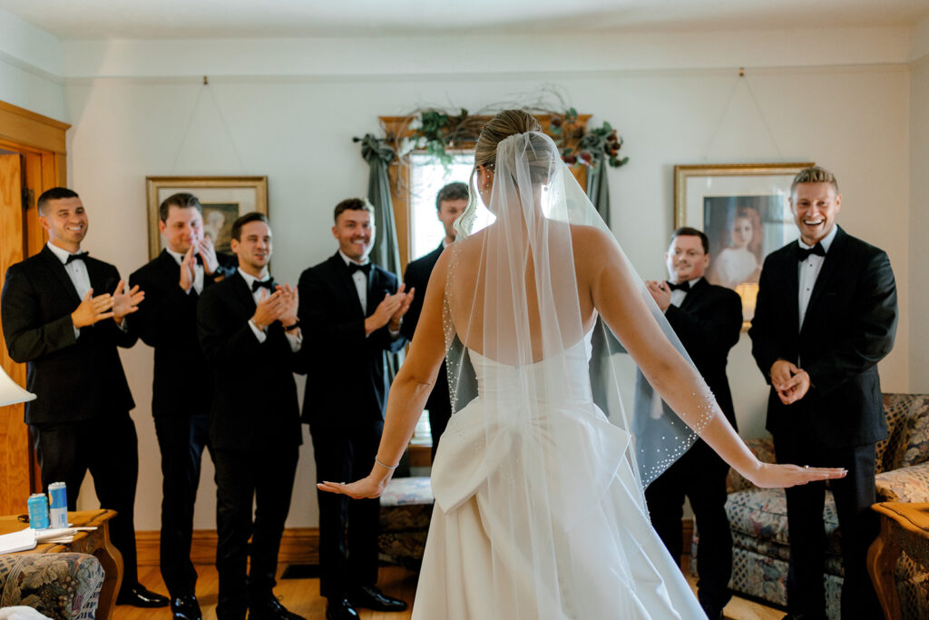 Bride surrounded by groomsmen reacting to her dress reveal, a playful pre-ceremony moment captured candidly.